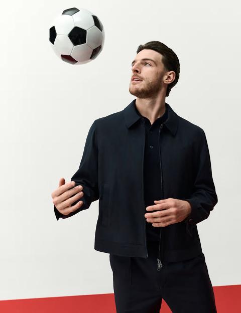 Declan Rice in a dark jacket, dark polo shirt, and dark trousers, looking up at a soccer ball.