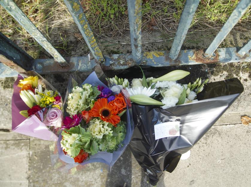 Three bouquets of flowers and a note rest by a rusty fence post, marking the scene of a fatal car crash.