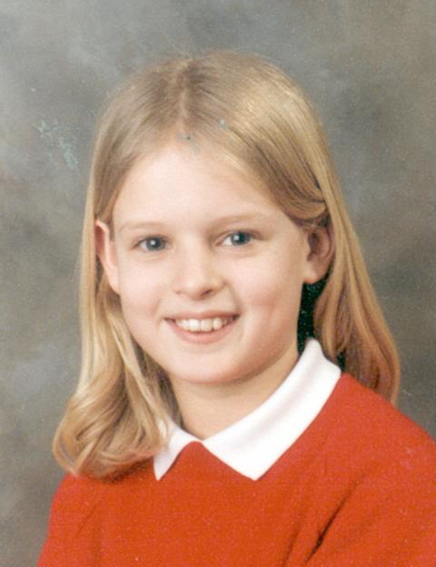 A young girl with blonde hair smiles in a school photo, wearing a red top with a white collar.