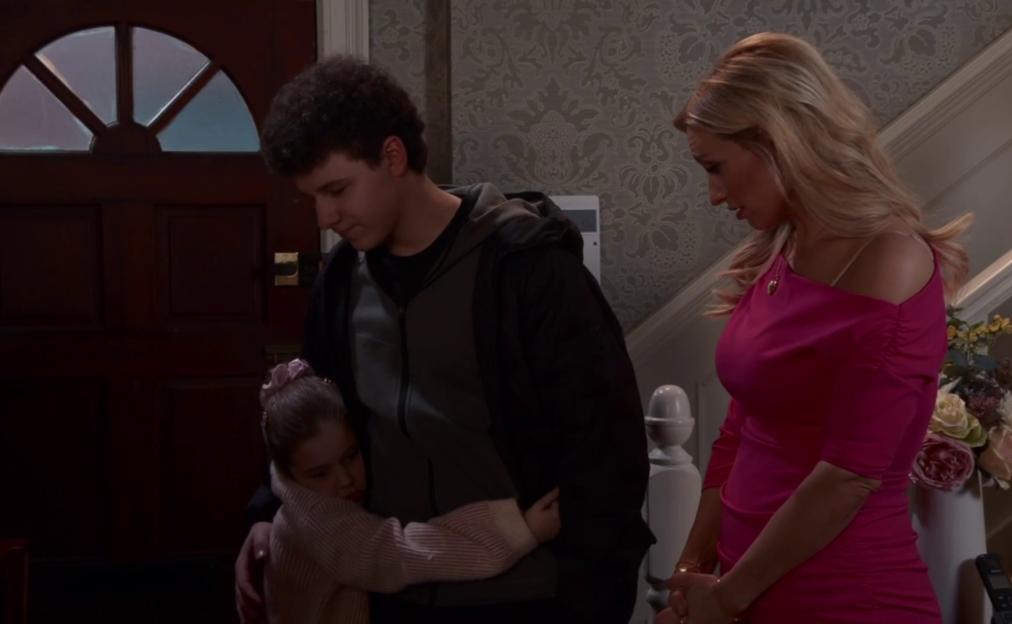 A young boy comforts a little girl while a woman in a pink dress looks on.