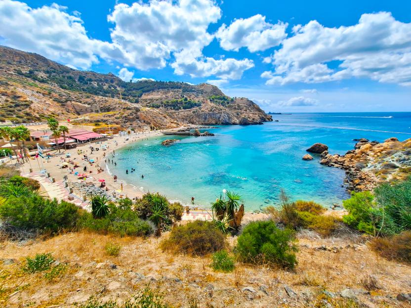 Cala Cortina beach in Cartagena, Spain, with people swimming and sunbathing in turquoise waters surrounded by rocky hills.