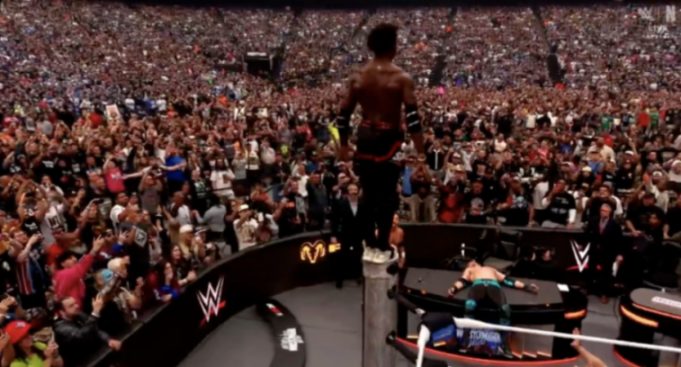 A shirtless wrestler stands on a ring post, facing a cheering crowd, while another wrestler lies prone on a table below.
