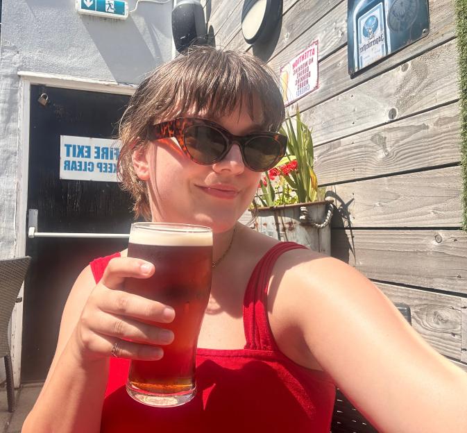 A woman in sunglasses and a red top holds a pint of beer outdoors at The Ardwick Pub.