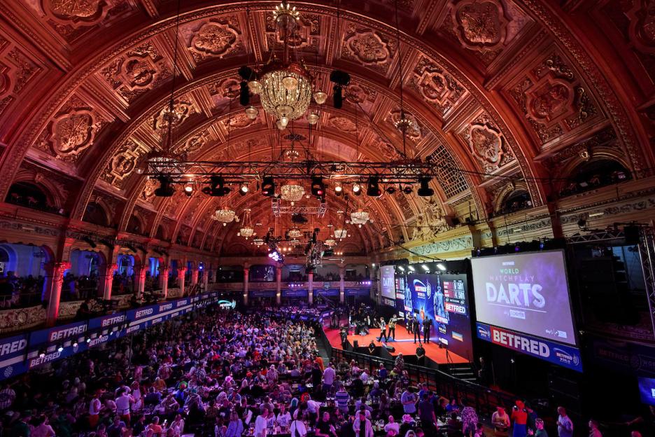 A large hall with a decorated arched ceiling, full of people watching a World Matchplay Darts tournament.