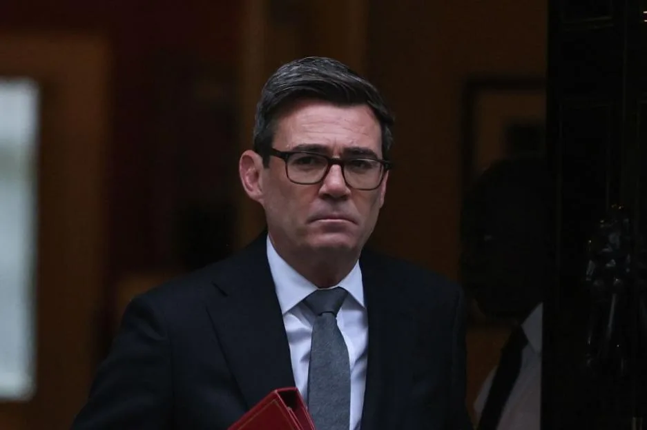 British Prime Minister Andy Burnham wearing a suit and glasses while holding a red folder.
