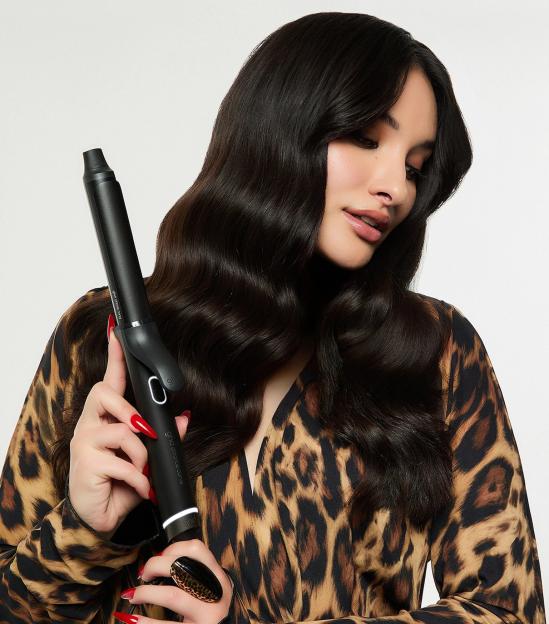 Woman with long dark wavy hair, wearing a leopard print dress, holding a ghd curling wand.