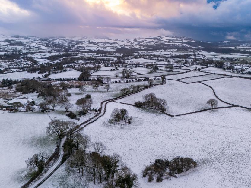 Aerial view of a snow-covered village and fields with trees, surrounded by hills under a cloudy sky.