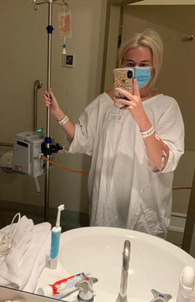 A woman in a hospital gown and mask takes a selfie in a bathroom while holding an IV pole.