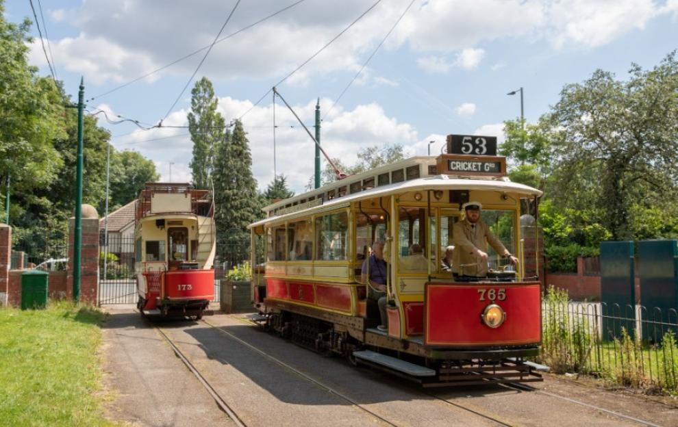 Two red and yellow Heaton Park trams, one with a driver in uniform, on tracks outdoors.