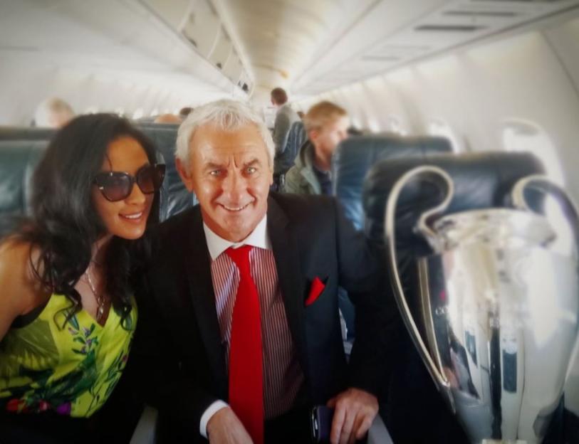 A man and woman smiling on an airplane next to the Champions League trophy.