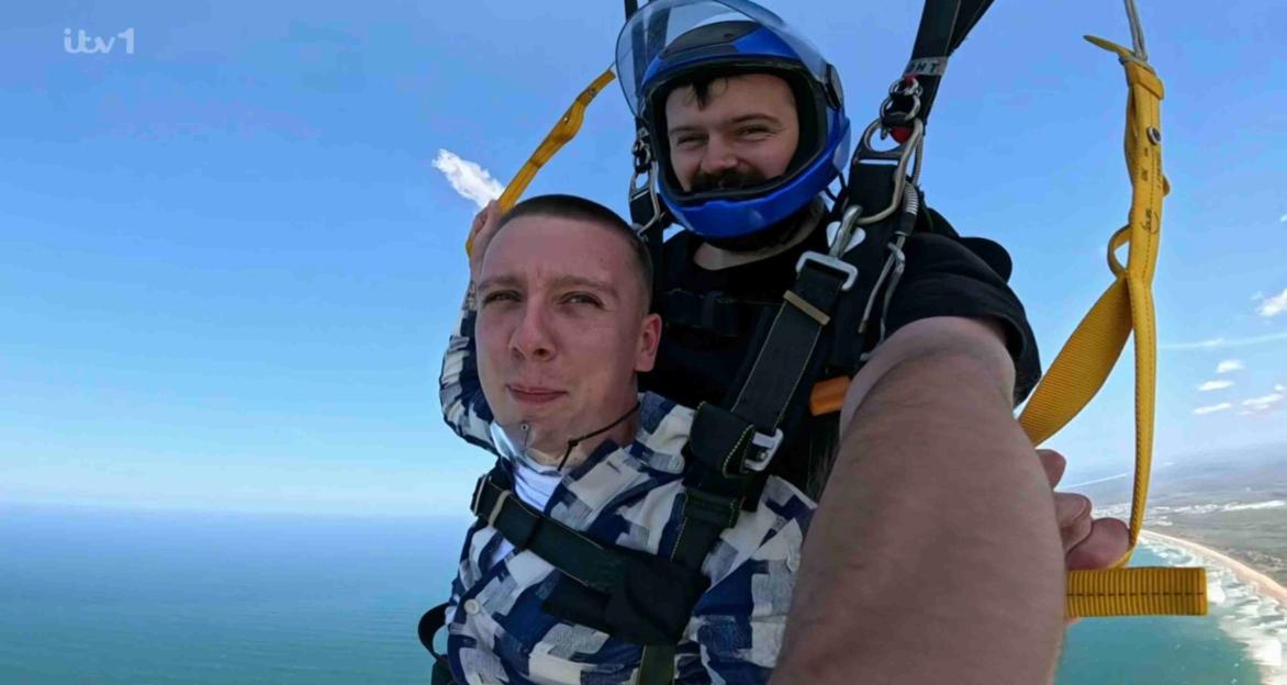 Aitch and another person skydiving, with a blue sky and ocean in the background.