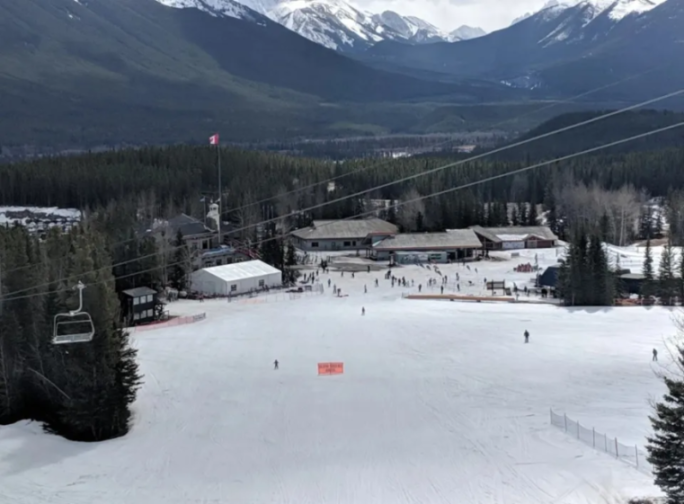 View of a ski resort from above, with buildings, a Canadian flag, skiers, and a ski lift visible.