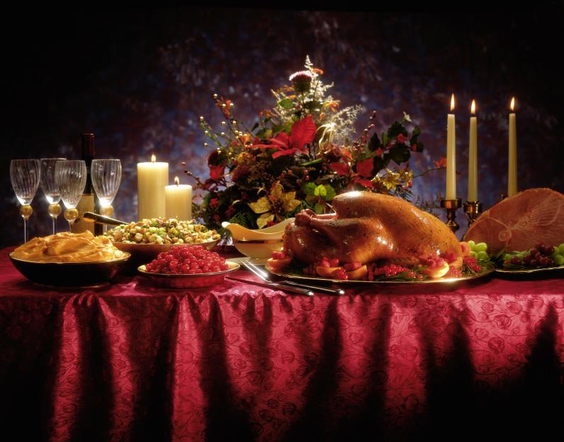 A holiday feast with turkey, ham, candied yams, stuffing, cranberry sauce, and a floral centerpiece on a red tablecloth.