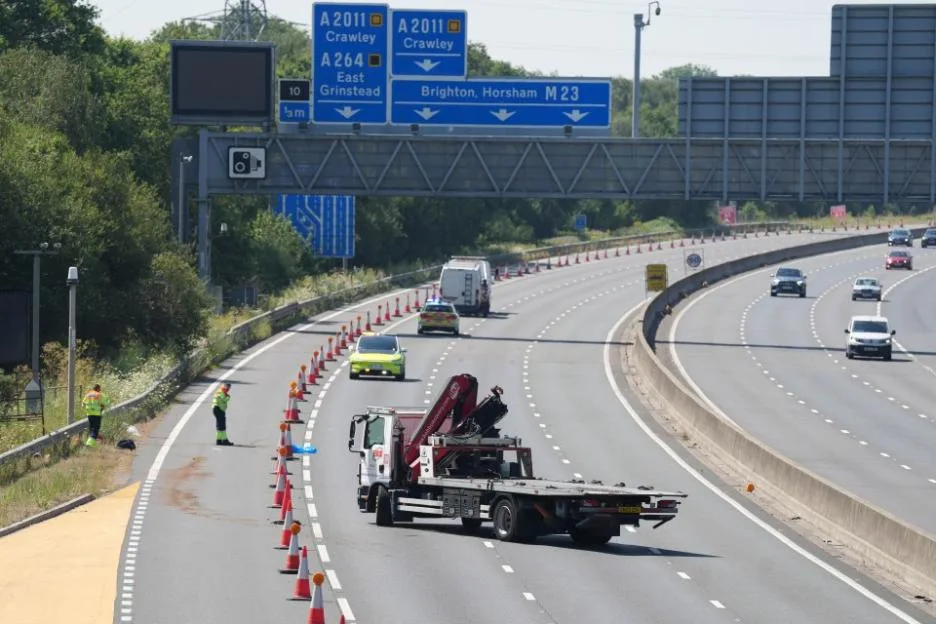 A flatbed truck and several vehicles on a divided highway, with cones marking off a lane where two workers are standing near the shoulder.