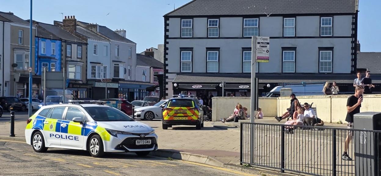 Two police cars are parked near a bus stop, with people sitting and standing nearby, and buildings lining the street.