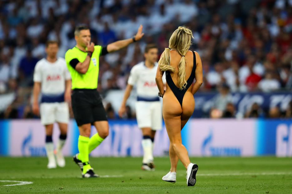 A streaker in a black swimsuit with her back to the camera walks across a soccer field as a referee blows a whistle and gestures with his hand for her to stop.