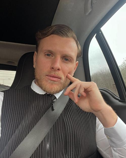 Tommy Mallet in a car, wearing a black pleated vest and white shirt, with a seatbelt across his chest.