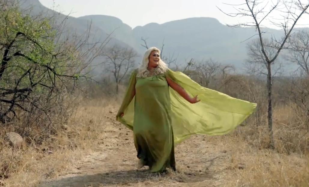 Gemma Collins in a flowing green dress with a cape walking in a dry, mountainous landscape.