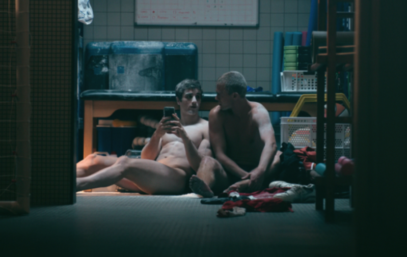 Two men sitting naked on the floor in a locker room, one looking at a smartphone.