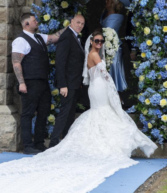 Tyson Fury in a tuxedo standing with a bride in a white wedding dress with a long train, with blue and yellow flowers behind them.