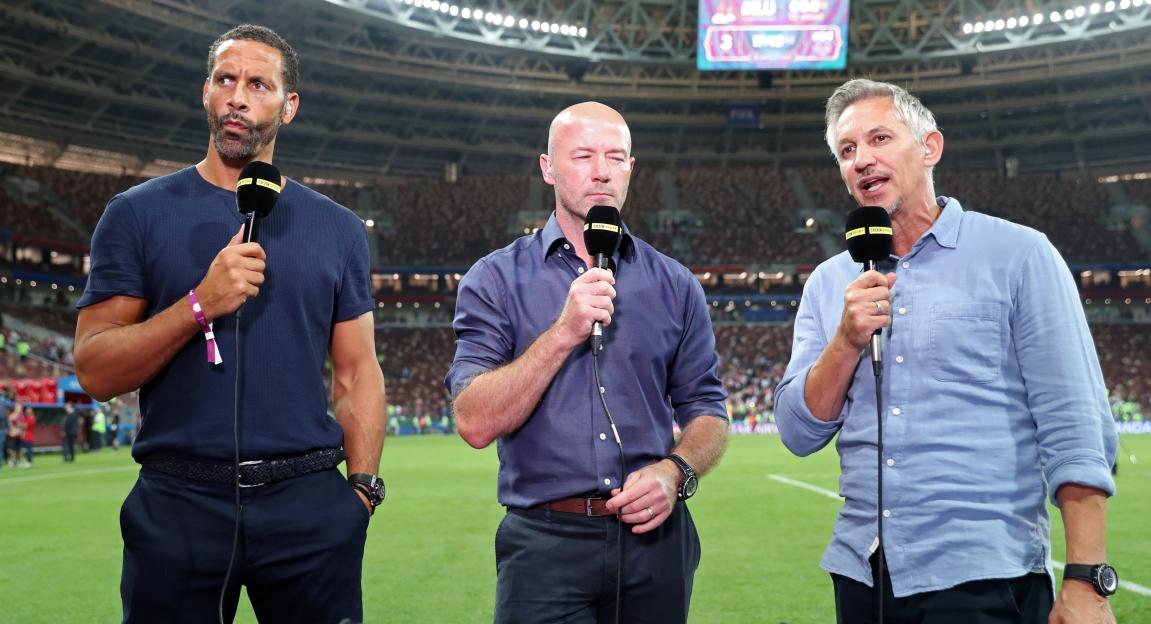 BBC Pundits Rio Ferdinand, Alan Shearer, and presenter Gary Lineker holding microphones at a football stadium.
