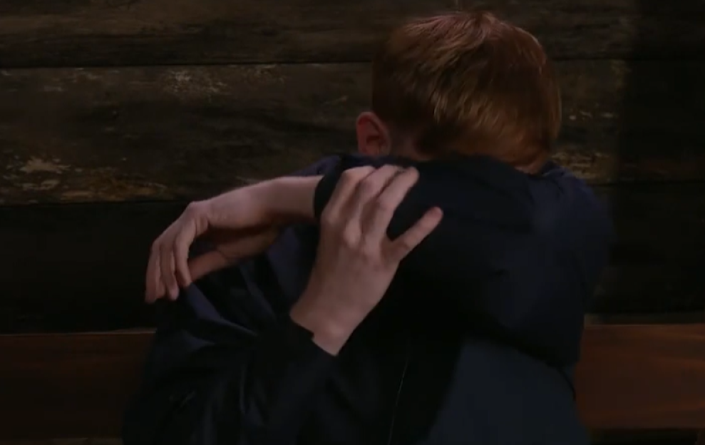 Two people in dark clothing embrace, one with red hair.