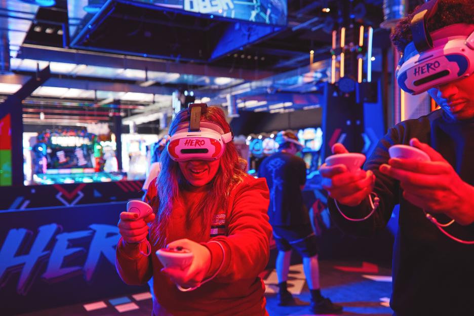 Two people wearing VR headsets and holding controllers, illuminated by red and blue light in an arcade.