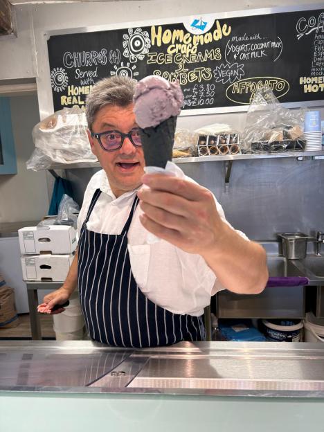 Ugur Vata, owner of The Gallery Creamery and Deli, holds up an unusual black garlic and peach ice cream in a black cone, smiling.