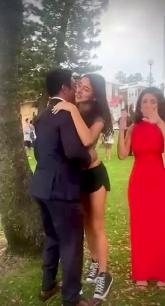 An engaged couple embracing with a woman in a red dress cheering in the background.