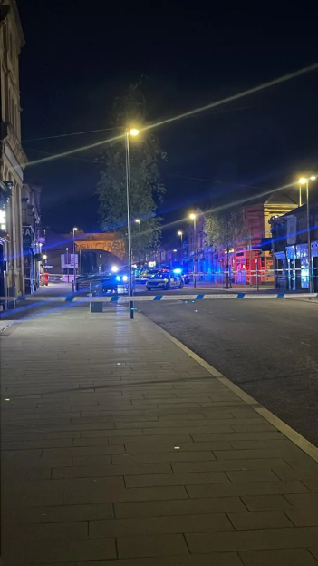 Police tape blocking off a city street with flashing blue police lights in the distance.