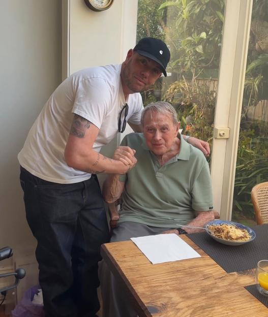 Terence Donovan, father of Jason Donovan, in a green polo shirt seated at a table, holding hands with a younger man in a white t-shirt and black cap, standing beside him.