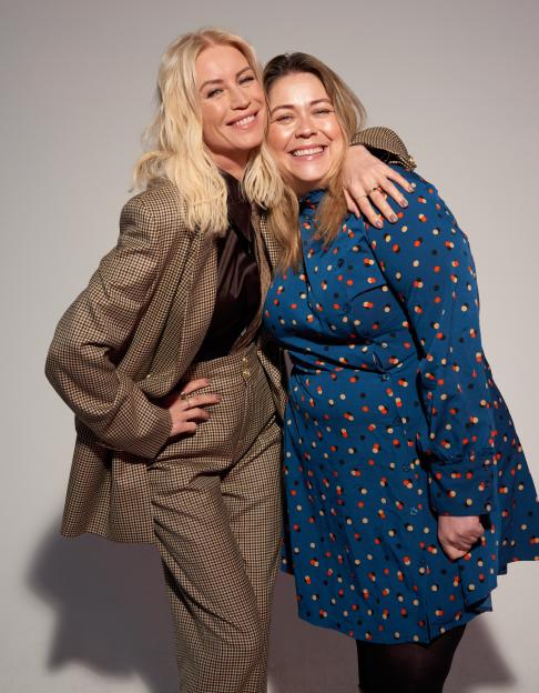 Two women smiling, one with blonde hair in a plaid suit, the other with brown hair in a blue polka-dotted dress, with her arm around the first woman.
