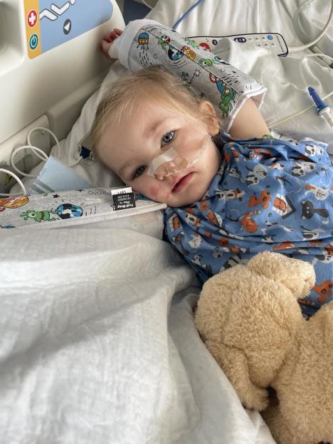 A young child with a nasal cannula lies in a hospital bed, wearing a blue patterned shirt and a cast on their arm.