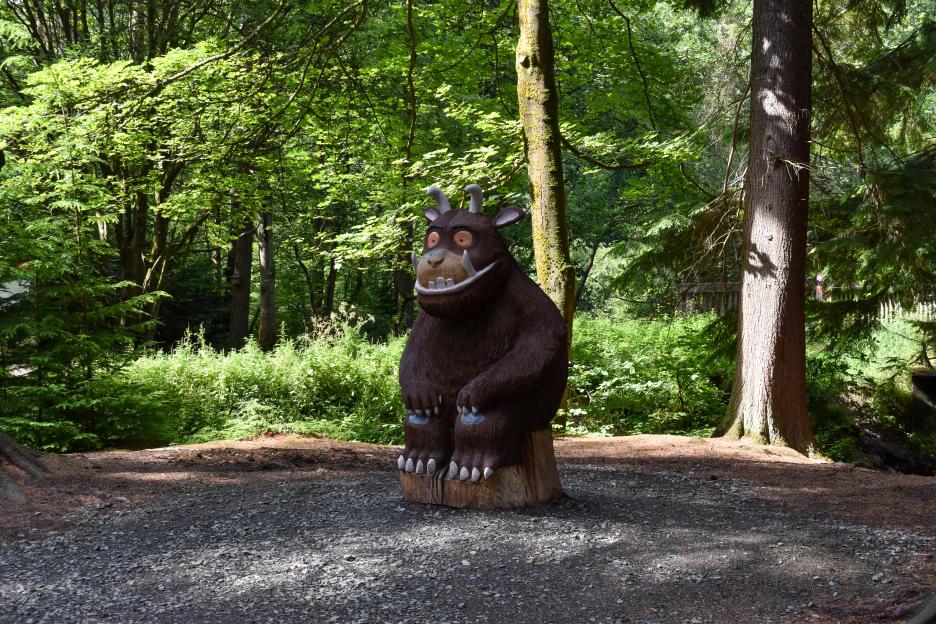 A wooden sculpture of the Gruffalo character sits on a tree stump in a forest.