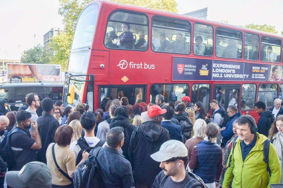 Crowds boarding a red double-decker First Bus, with people visible inside the bus and in the crowded street.