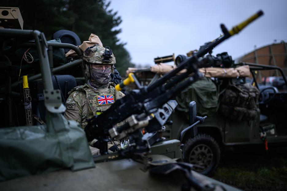 A Special Operations Land Task Group member in camouflage uniform and balaclava sitting in a military vehicle with a Union Jack flag on their chest.