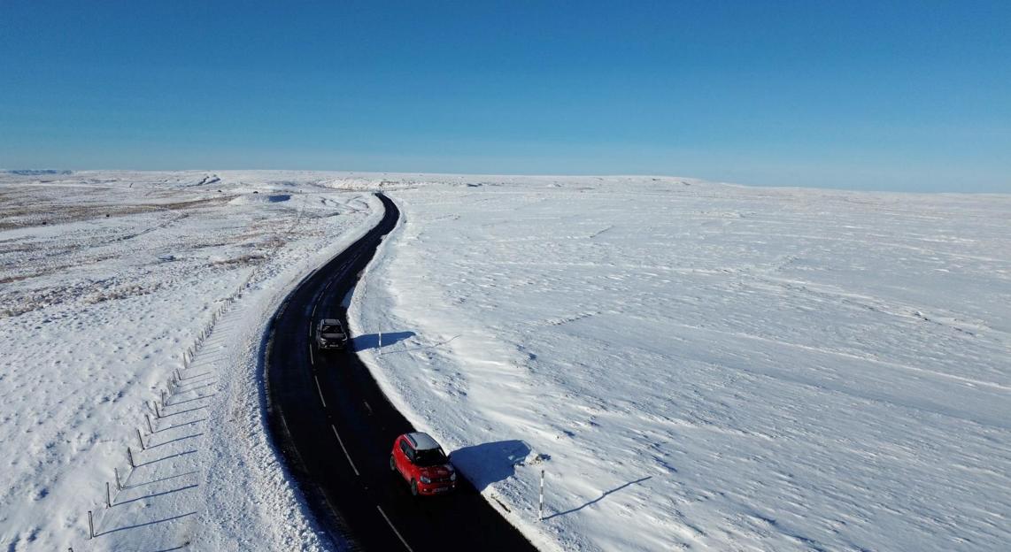An aerial view of a winding road through a snow-covered moor with two cars on it.