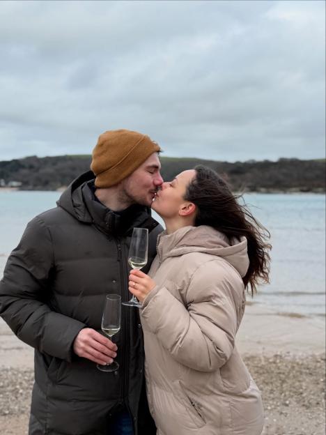 Evie Pickerill and Alex Phelps kiss while holding champagne flutes, showcasing an engagement ring on Pickerill's finger, on a beach.