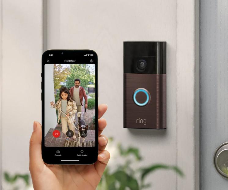 Hand holding a smartphone displaying a live video feed from a Ring doorbell, showing a man, a girl, and a dog at the front door.