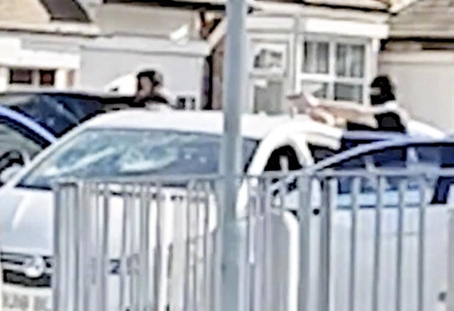 A man in a black mask standing out of a car window, holding a gun, and pointing it at another person across the street.