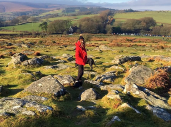 A person in a red jacket and a black dog stand on a rocky, grassy hill overlooking a valley with trees and a long line of parked cars in the distance.