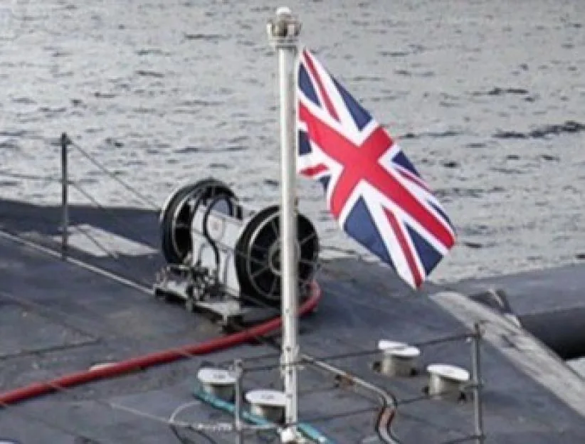 A Royal Navy submarine moored at HM Naval Base Clyde in Faslane, Scotland, with a large industrial building and a crane in the background.
