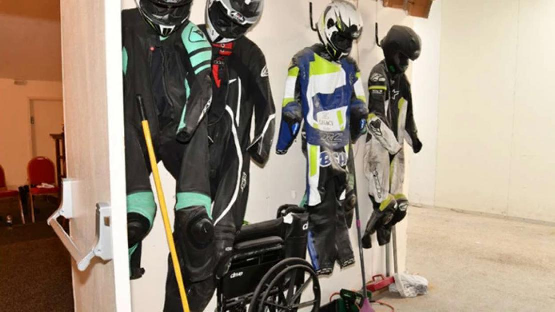 Motorcycle racing suits and helmets hanging on a wall next to a wheelchair.