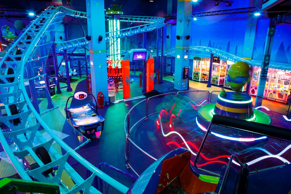 Amusement park with a blue-lit roller coaster, brightly lit arcade games, and a rotating spinning ride.