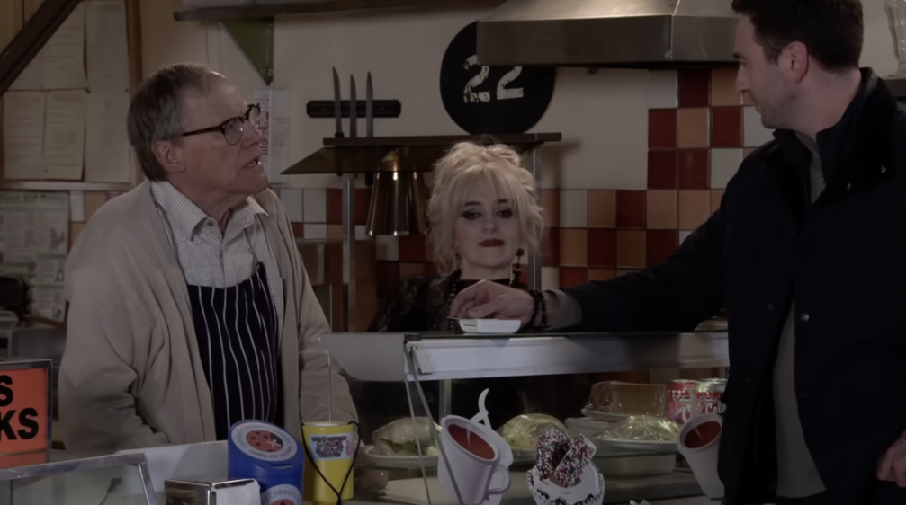 Three people interacting behind a counter with food items, including salads, cakes, and drinks.