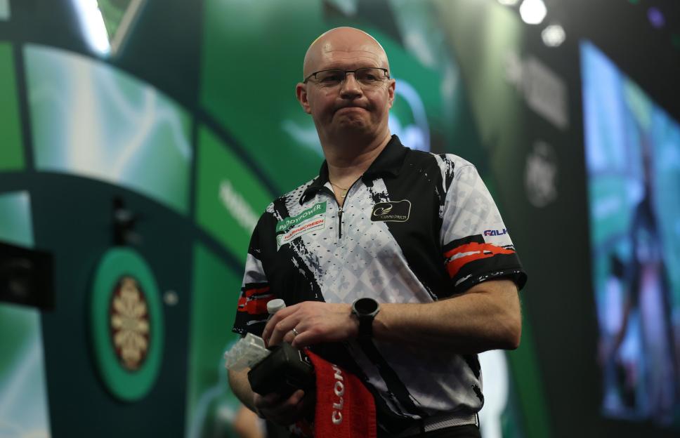 A bald man in glasses with a concerned expression, wearing a black, white, and red dart shirt, holds a water bottle and a red towel with a white logo, while standing near a dartboard at the Paddy Power World Darts Championship.