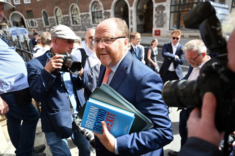 A man in a suit carrying a book titled "Britain Beyond Brexit" surrounded by photographers.