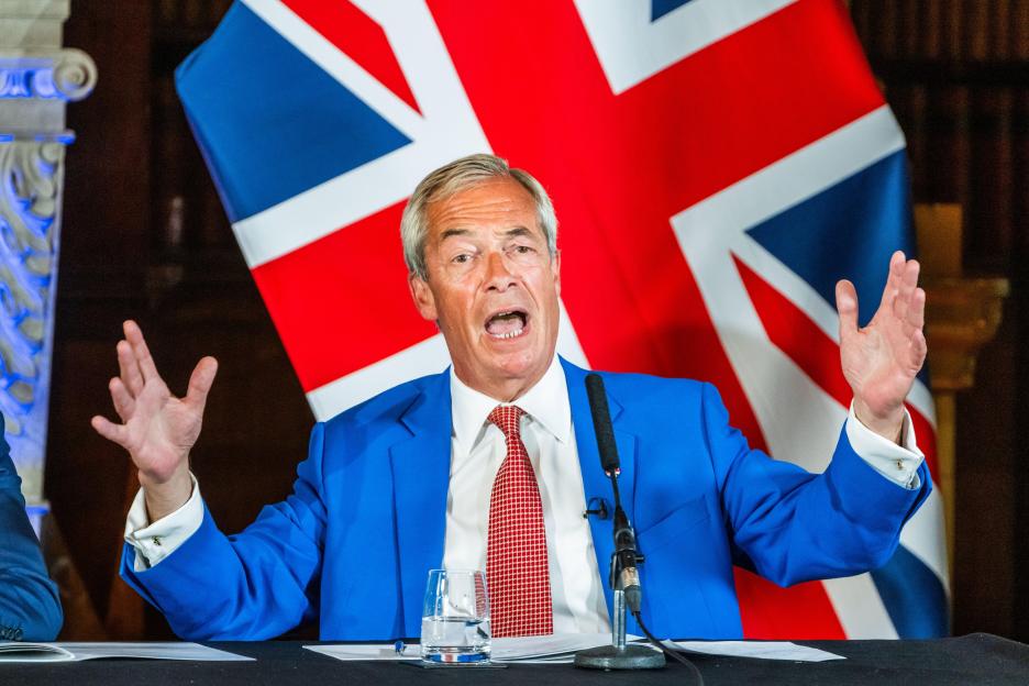 Nigel Farage speaking at a press conference with the Union Jack flag behind him.