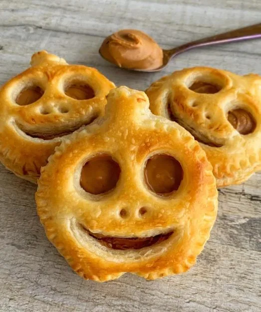 Three Biscoff puff pastry pumpkins with carved jack-o'-lantern faces, filled with Biscoff spread, and a spoon of spread in the background.