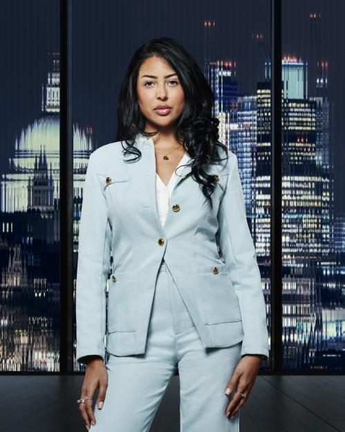 Pascha Myhill, a contestant on The Apprentice, poses in a light blue pantsuit in front of a window overlooking a cityscape at night.